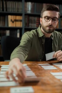 A focused young man sorting cards on a table in a library, surrounded by bookshelves.