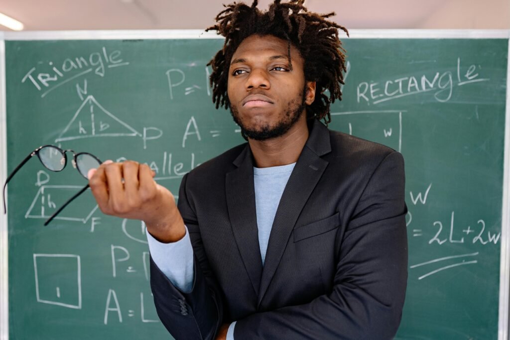 Educator posing with eyeglasses in hand, chalkboard filled with geometric formulas.